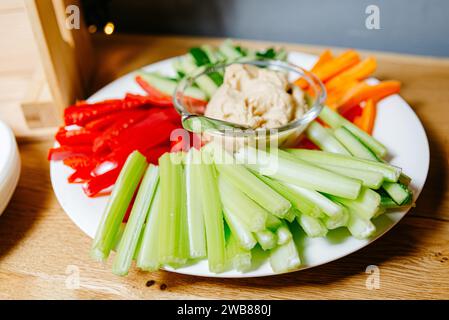 Farbenfrohe Gemüse-Snackplatte mit frischen Selleriestangen, Karottenstäbchen, rotem Paprika und cremigem Hummus-Dip in einer Glasschale. Stockfoto