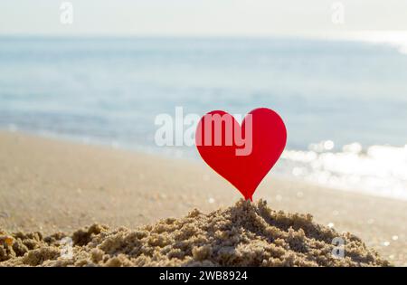 Plastikstab in Form eines roten Herzens im Sand auf Sandstrand der Küste auf Hintergrund der Meereswellen an sonnigem Sommertag Nahaufnahme. Konzept Liebe Beziehung Romantik Amour St. Valentinstag Stockfoto