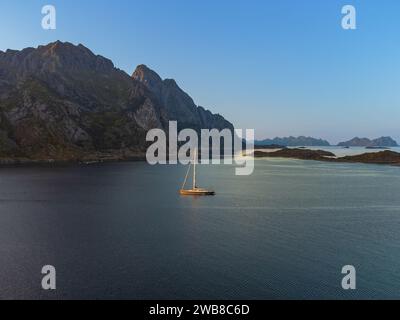 Ein Segelboot unter der Mitternachtssonne, hinter einem großen Hügel in Lofoten, bei Henningsvaer, in der Ferne hohe Berge, ruhiges Meer. Stockfoto