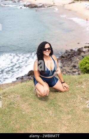 Schöne, lockige Frau mit Sonnenbrille, die sich auf einem Hügel vor Meer und Felsen im Hintergrund hockt. Reisende Person. Stockfoto