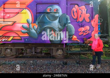 As, Limburg, Belgien. Dezember 2023. Güterwagen mit Graffiti bemalt, Senior Tourist posiert mit seinem Dackel, Roboter auf lila Hintergrund, Disu Stockfoto