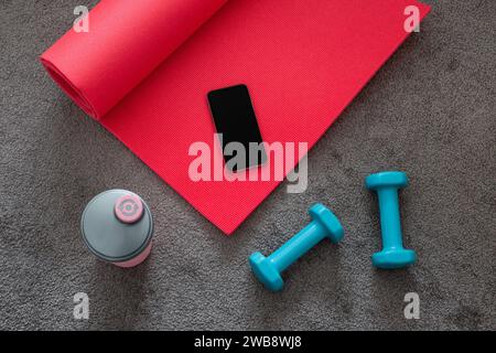 Blick von oben auf rosafarbene Yogamatte, blaue Kurzhanteln, Fitnessgeräte auf grauem Teppich mit Mobiltelefon. Stockfoto