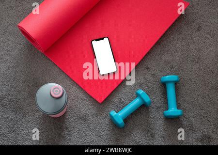 Blick von oben auf rosafarbene Yogamatte, blaue Kurzhanteln, Fitnessgeräte auf grauem Teppich mit Mobiltelefon. Stockfoto