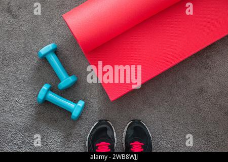 Blick von oben auf rosafarbene Yogamatte, blaue Kurzhanteln, Fitnessgeräte auf grauem Teppich mit Schuhen. Stockfoto