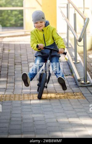 Ein kleiner Junge fährt mit seinem Balance Bike bergab, hebt beide Beine und lernt, zu balancieren. Stockfoto