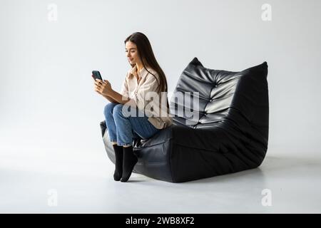 Foto-Porträt Ganzkörperansicht eines lächelnden Mädchens, das Telefon in zwei Händen hält und im Sitzsack-Stuhl sitzt Stockfoto