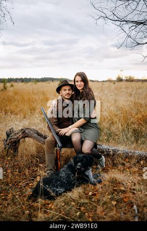 Das junge, reizende Paar, der Jäger und seine Dame sitzen nebeneinander mit ihrem Hund in der Natur Stockfoto