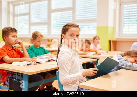 Seitliches Porträt eines Schulmädchens, das Buch hält, während er mit Freunden im Klassenzimmer auf der Bank sitzt Stockfoto