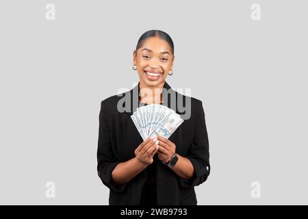 Lächelnde afroamerikanische Geschäftsfrau in einem stilvollen schwarzen Blazer, die einen Geldfan freudig hält Stockfoto