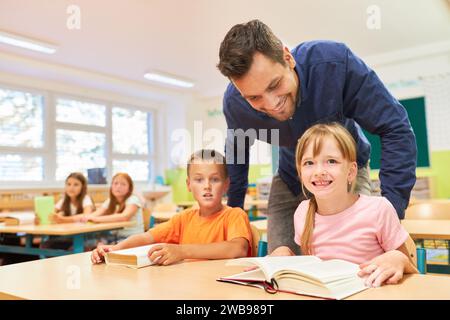 Glücklicher männlicher Lehrer, der neben Schülern steht und Bücher liest, während er im Klassenzimmer auf der Bank sitzt Stockfoto