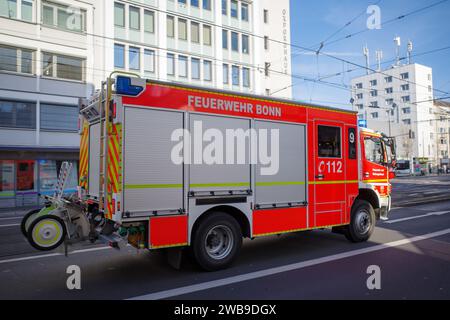 Roter Feuerwehrwagen der Feuerwehr Bonn in Bewegung auf einer Stadtstraße mit Gebäuden im Hintergrund, blinkende Lichter auf leerer Straße. Stockfoto