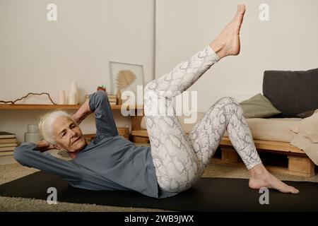 Aktive Rentnerin in Sportbekleidung, die im Schlafzimmer auf einer Matte liegt und mit einem Bein und den Händen auf dem Hinterkopf körperliche Bewegung macht Stockfoto