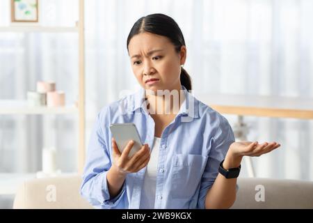 Verärgerte chinesin, die auf der Couch sitzt, auf das Telefon schaut, Gesten macht Stockfoto
