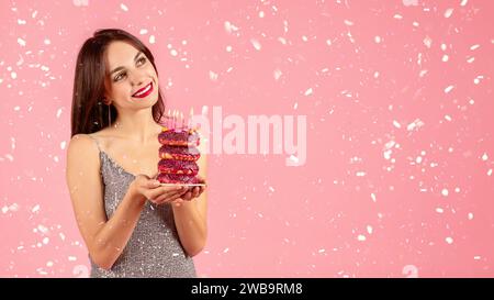 Lächelnde Frau in einem glitzernden Kleid, die einen Stapel rosafarbener Donuts bewundert, die mit Geburtstag geschmückt sind Stockfoto