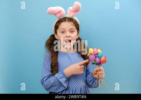Porträt eines charmanten fröhlichen Mädchenkindes in osterhasenohren, der auf kleine farbige Eier zeigt, Ausdruck Genießen Sie die Feier des christlichen Urlaubs Stockfoto