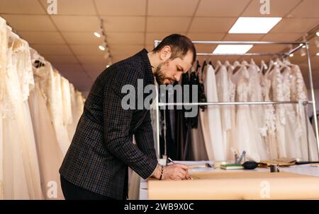 Junger seriöser Schneider mit Bart, der auf der Zeichnung neben einem Holztisch mit Fäden in einem fantastischen Atelier mit antiken Möbeln und Schaufensterpuppe blickt Stockfoto