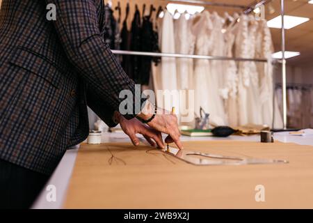 Junger, seriöser Schneider mit Bart, der in der Nähe eines Holztisches mit Fäden in einem fantastischen Atelier mit antiken Möbeln und Schaufensterpuppe zeichnet Stockfoto