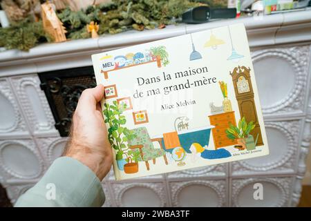 Paris, Frankreich - 16. Dezember 2023: Männliche Hand präsentiert Dans la maison de mA Grand-mere von Alice Melvin, ein Kinderbuch, vor einem rustikalen Ofen Stockfoto