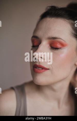 Porträt einer Frau mit geschlossenen Augen, die ein außergewöhnliches Make-up mit roten Lidschatten und rotem Lippenstift trägt, mit festgebundenen Haaren Stockfoto