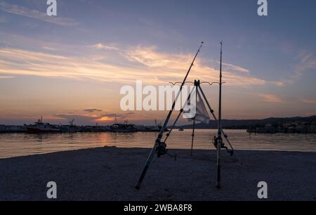 Angelruten stehen am Rand eines Piers in einer Bucht mit Fischerbooten bei Sonnenuntergang in Skioni, Griechenland Stockfoto