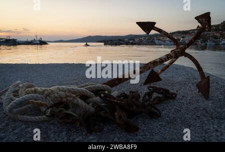 Ein alter rostiger Anker mit einem Seil liegt am Rand eines Piers in einer Bucht mit Fischerbooten in Skioni, Griechenland Stockfoto