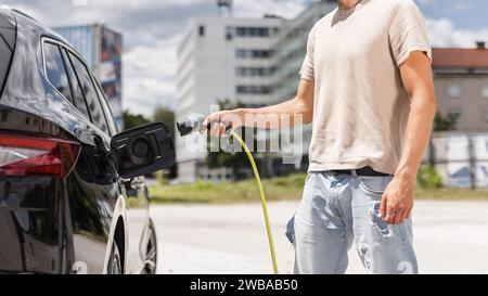 Mann, der eine Steckdose öffnet und ein Ladegerät in ein schwarzes Elektroauto an eine EV-Ladestation in der Stadtstraße stellt Stockfoto