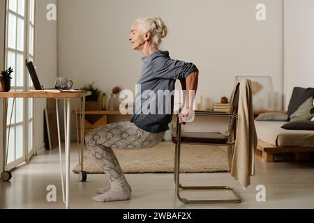 Seitenansicht einer älteren aktiven Frau in Freizeitkleidung, die Squats vor dem Laptop macht, während sie am Stuhl hält und während des Trainings auf den Bildschirm blickt Stockfoto