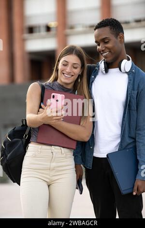 Multirassisches Paar steht und sieht sich einen Anruf auf dem Campus an. Zwei junge Studenten machen ein Selfie und lächeln auf der Straße. Stockfoto