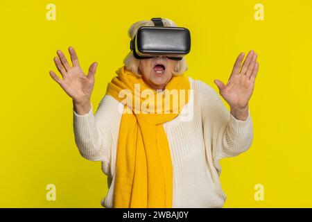 Begeisterte, glückliche Seniorin, die die Headset-Helm-App nutzt, um ein Simulationsspiel zu spielen. 3D 360-Videos in Virtual Reality ansehen. Reife Großmutter in VR-Brille isoliert auf gelbem Hintergrund. Zukunftstechnologie Stockfoto