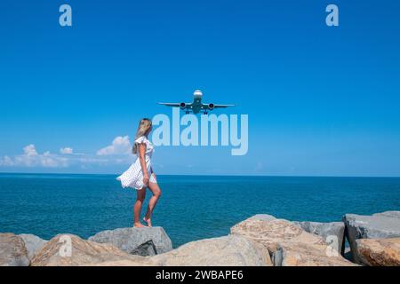 Seaside Fantasy: Mädchen in weißem Kleid, Steine, Blaues Meer und ein fliegendes Flugzeug Stockfoto