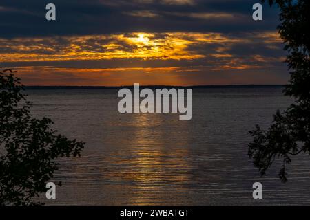 Wunderschöner Sonnenuntergang oder Sonnenaufgang mit orangefarbener Sonne, die durch dunkle Wolken über dem Horizont scheint und sich auf dem Seewasser reflektiert. Stockfoto