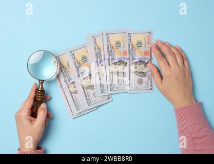 100 Dollar-Scheine und eine Frauenhand, die eine Lupe auf blauem Hintergrund hält, Draufsicht Stockfoto