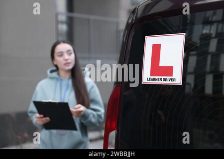 Kursleiter mit Klemmbrett in der Nähe des Autos im Freien, selektiver Fokus auf L-Platte. Fahrschule Stockfoto