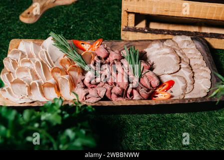 Ein rustikales hölzernes Servierbrett mit dünn geschnittenem Roastbeef und truthahn, garniert mit frischem Rosmarin und Chili-Pfeffer, präsentiert auf Kunstrasen. Stockfoto