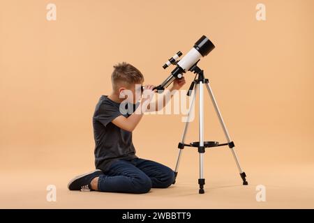 Kleiner Junge, der Sterne durch ein Teleskop auf beigefarbenem Hintergrund ansieht Stockfoto