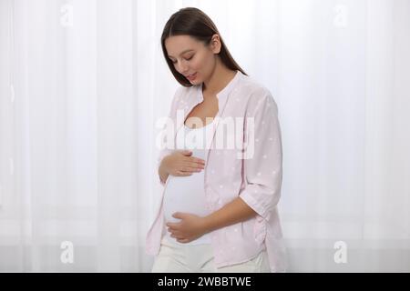 Schöne schwangere Frau in rosa Hemd in der Nähe des Fensters drinnen Stockfoto