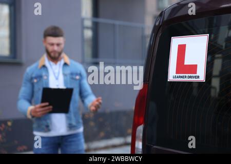 Kursleiter mit Klemmbrett in der Nähe des Autos im Freien, selektiver Fokus auf L-Platte. Fahrschule Stockfoto
