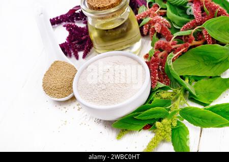 Amaranth Mehl in einer Schüssel, Samen in einem Löffel und Öl in einem Glas, braune, grüne und lila Blüten einer Pflanze auf weißem Holzbrett Hintergrund Stockfoto