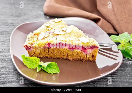 Ein Stück süßer Kuchen mit Himbeeren, Butterbrösel und Mandelflocken in Teller, Minze und einem Handtuch auf Holzbrett Hintergrund Stockfoto