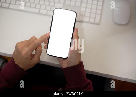 Draufsicht eines Mannes, der sein Smartphone an ihrem Schreibtisch verwendet. Ein Smartphone-Modell mit weißem Bildschirm in der Hand eines Mannes. Menschen- und Technologiekonzepte Stockfoto
