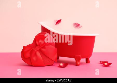 Mini-Badewanne mit Seifenschaum, Rosenblättern und Geschenkbox auf farbigem Hintergrund. Valentinstag-Feier Stockfoto