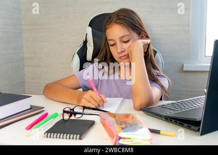 Kindererziehung. Gelangweilter Schüler müde vom Lernen, Hausaufgaben mit pc zu machen, trauriges Teenagermädchen, das auf die Prüfung vorbereitet, im Copybook d schreibt Stockfoto