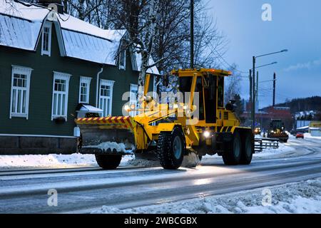 Gelber Motorgrader Veekmas FG 2327 S entfernt morgens Schnee von der Straße, ein weiterer Motorgrader folgt. Salo, Finnland. Dezember 2023. Stockfoto