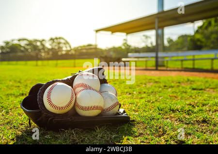 Baseballhandschuh gefüllt mit Baseballbällen, der während eines Sonnenuntergangs auf dem Ballfeld vor einer leeren Ausbuchtung sitzt Stockfoto