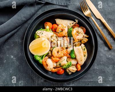 Gemischte Meeresfrüchte enthalten Garnelen, Muscheln, Calamari-Tintenfische und Fisch mit Brokkoli, Kirschtomate, Lemon on on Black Gericht auf Restauranttisch. Salat mit Meeresfrüchten auf dunklem Hintergrund. Draufsicht Stockfoto