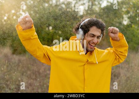 Junger tanzender Mann im gelben Regenmantel, der den Sommerregen genießt, während er im Park steht und Musik über Kopfhörer hört Stockfoto