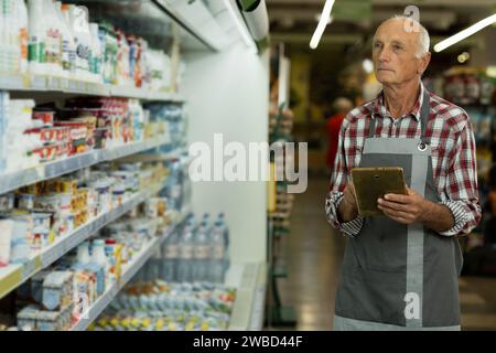 Ein Supermarktarbeiter auf einem Vorfeld, der in der Nähe von Regalen mit Milchprodukten steht und ein digitales Tablet verwendet, um Nachzählungen durchzuführen Stockfoto