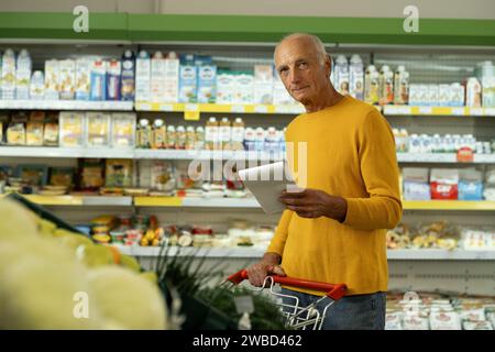 Senior Kundin überprüft ihre Einkaufsliste, während sie mit einem Wagen durch den Supermarkt geht, sich die Kamera anschaut, Käufer im Einzelhandel verkauft Stockfoto