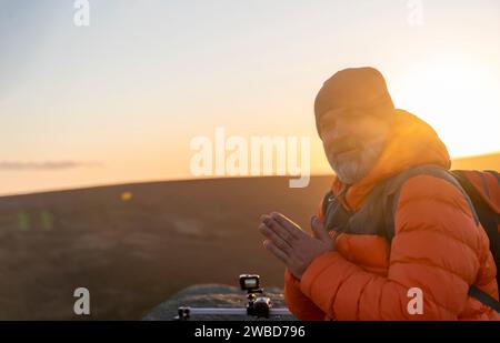 Bärtiger Mann in orangefarbener Jacke, der sich allein auf dem Gipfel des Berges entspannt und bei Sonnenaufgang heißen Kaffee trinkt. Travel Lifestyle Konzept der Nationalpark Stockfoto