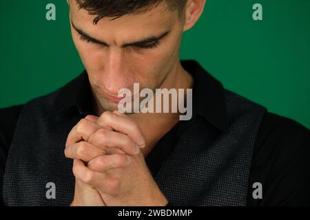 Ein junger Mann mit gefalteten Händen, der in Nahaufnahme betet. Emotionen eines gutaussehenden Mannes auf grünem Hintergrund, Chromakey-Nahaufnahme, dunkles Haar, junger Mann Stockfoto